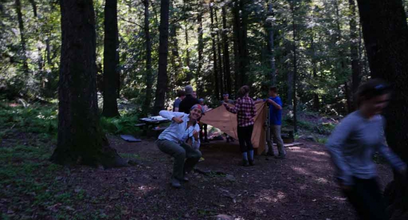 A group of students work to set up camp in a wooded area. A person in the foreground is giving the camera a thumbs up.